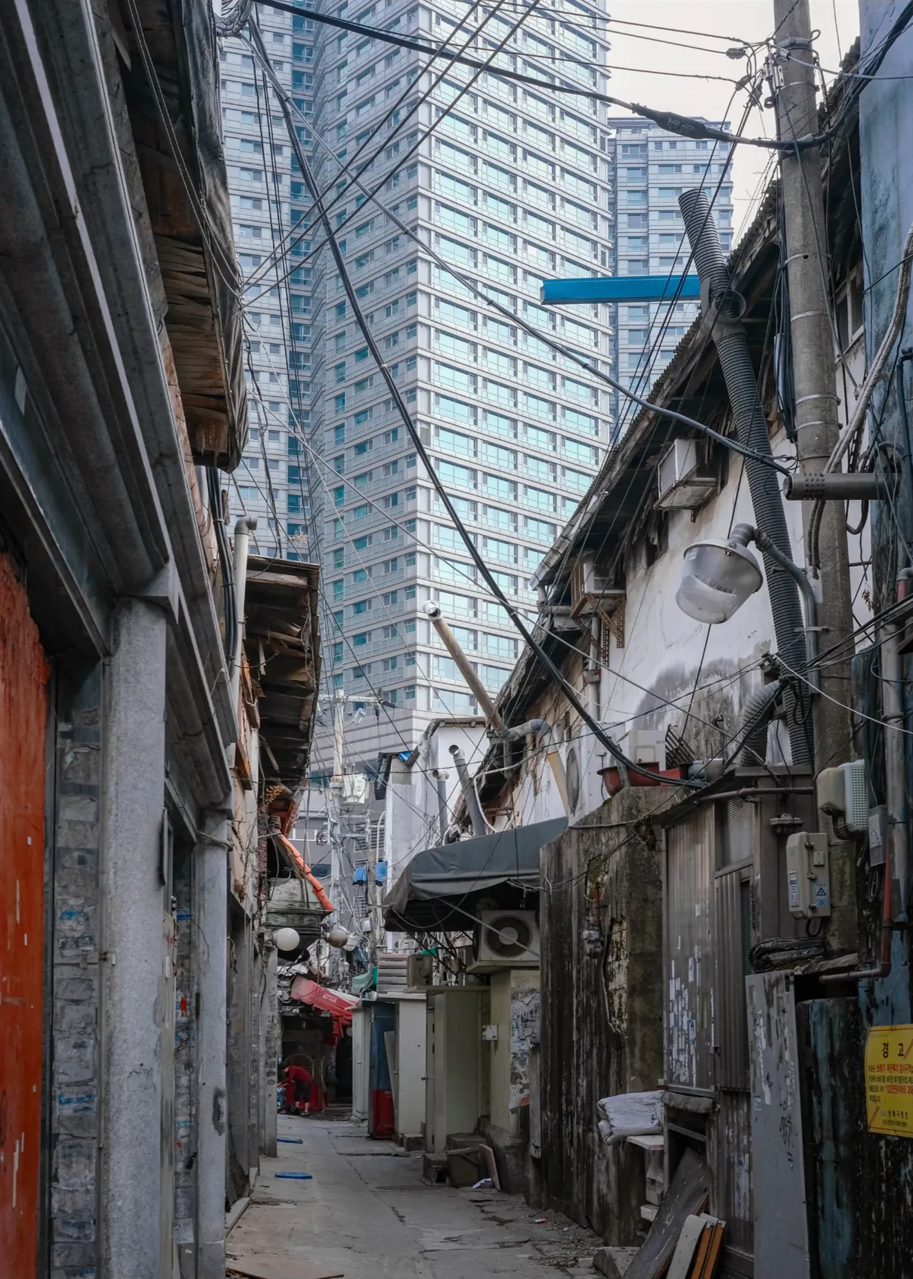 A narrow alley of old shopfronts and tangled wires, a glass tower rising behind