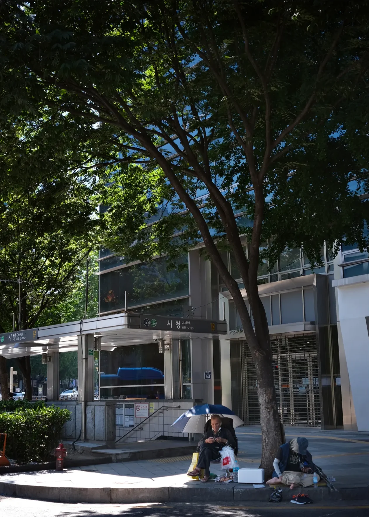 Two people resting under a tree beside the City Hall subway entrance, one under an umbrella