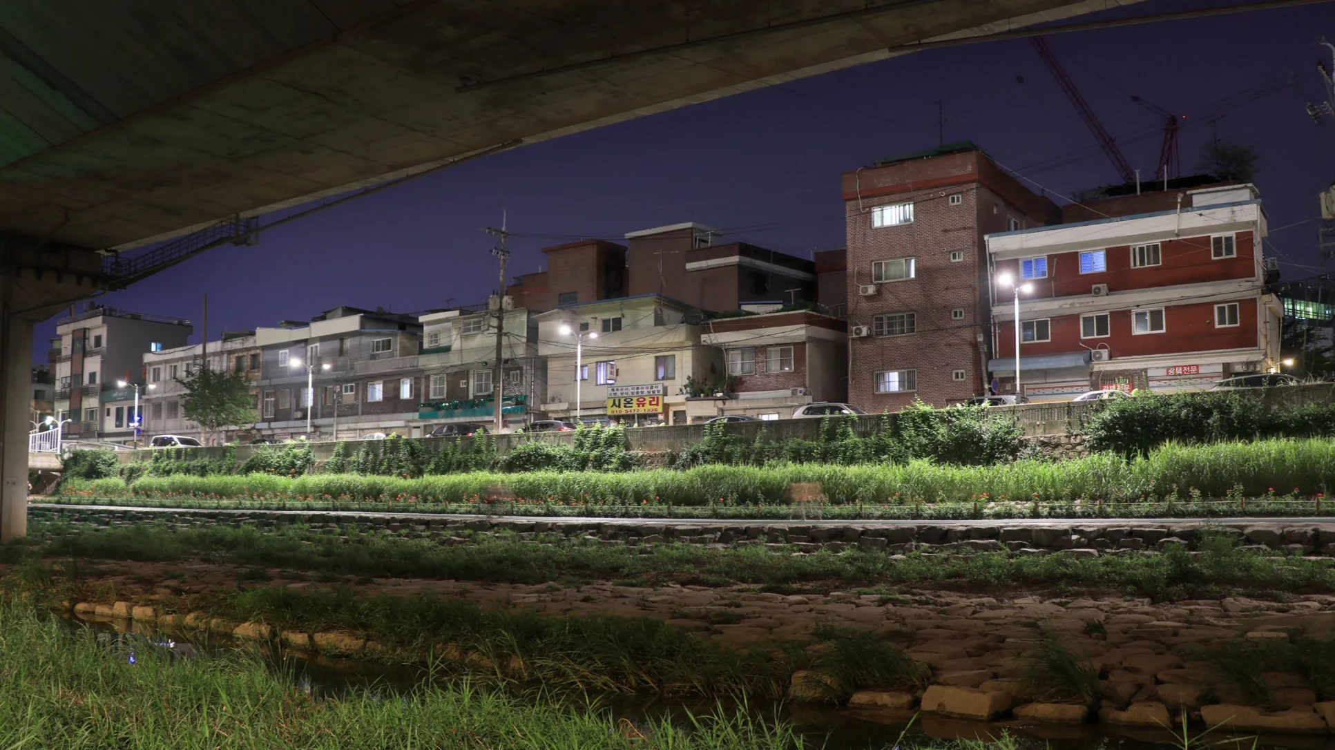 Low brick apartment blocks at dusk, seen from a stream bed beneath an overpass