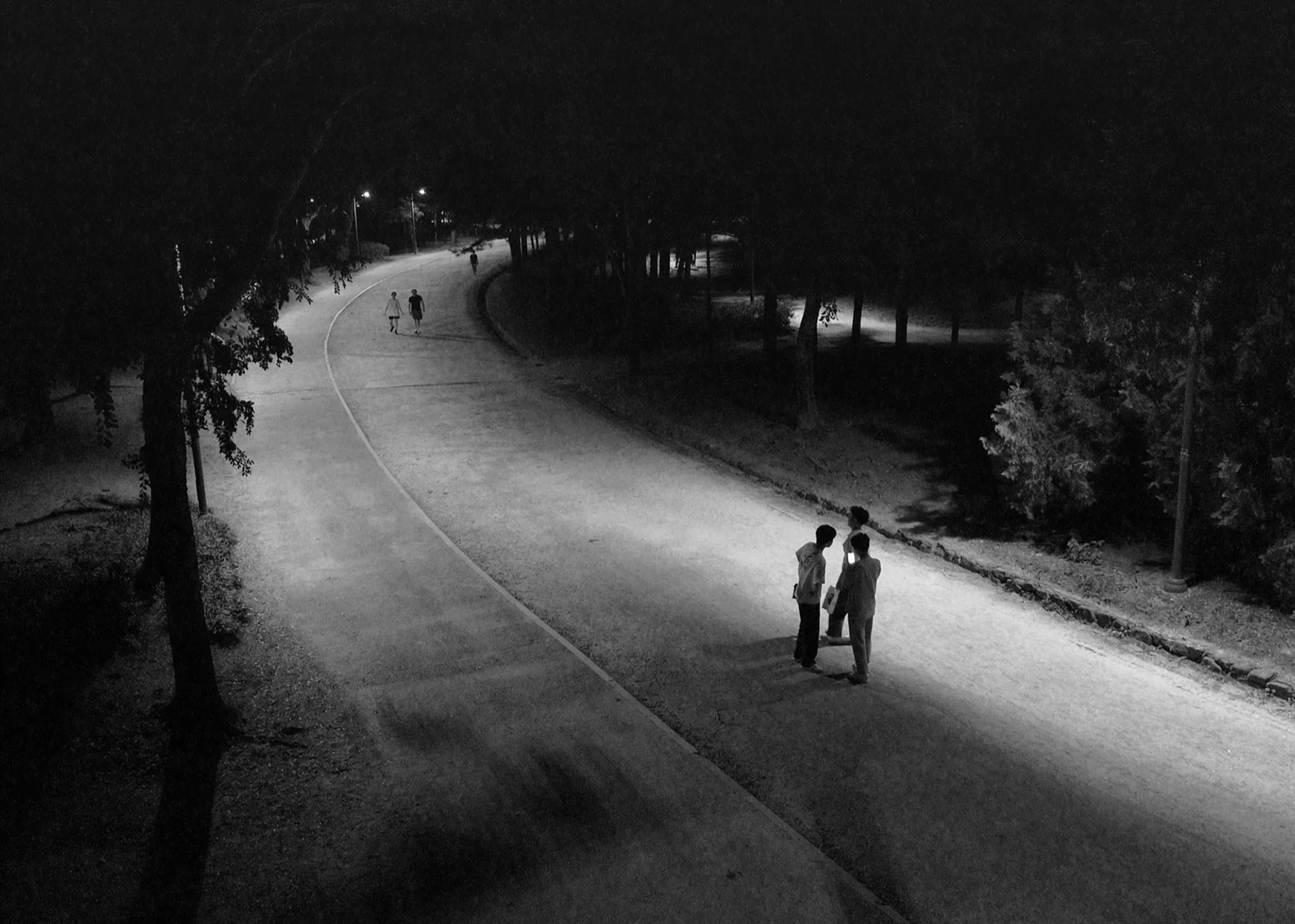 Black and white: a curving park path at night, three figures standing in a pool of lamplight