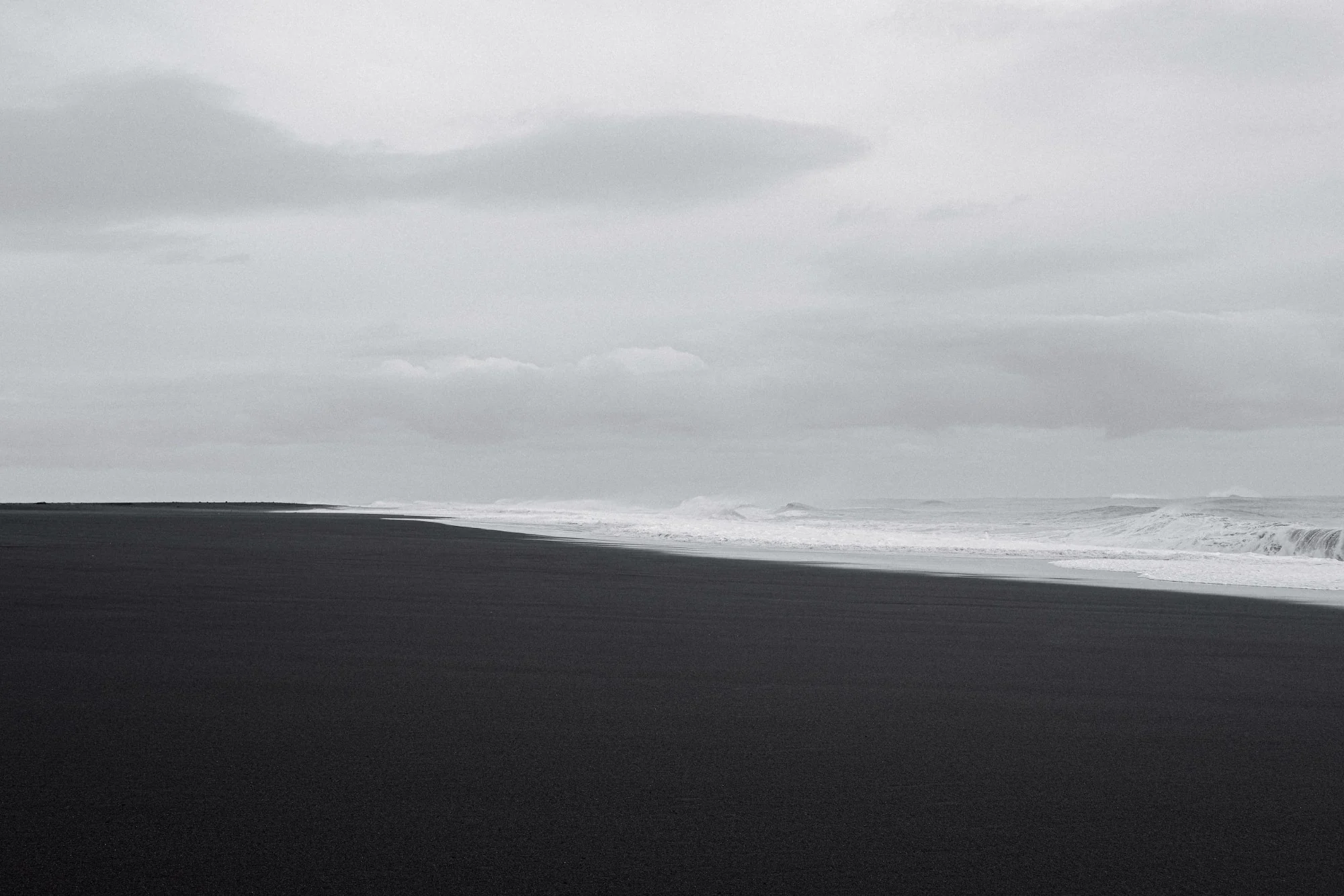Black and white: an empty black-sand beach under low cloud, surf breaking along the shore