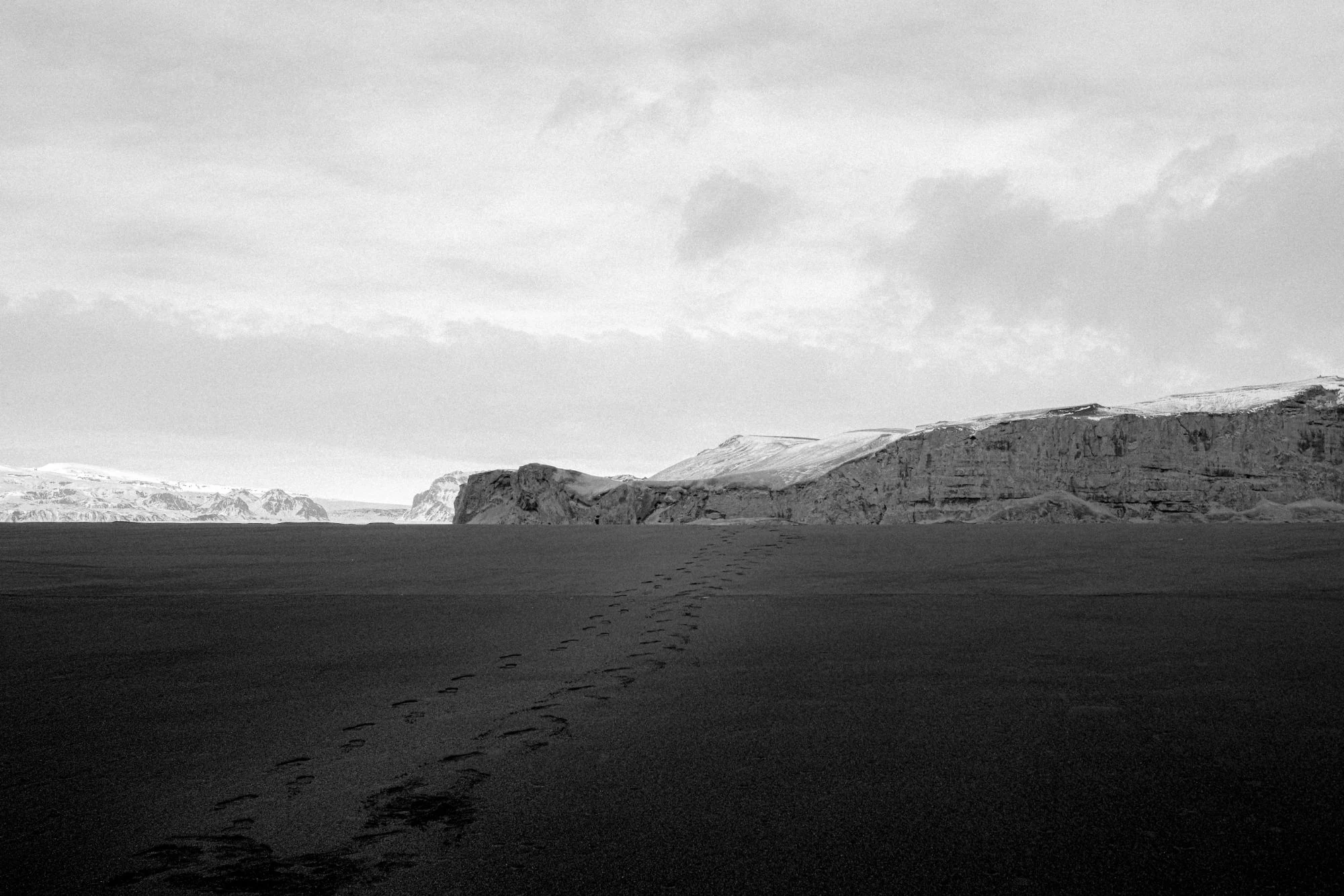 Black and white: a line of footprints across black sand toward snow-dusted cliffs