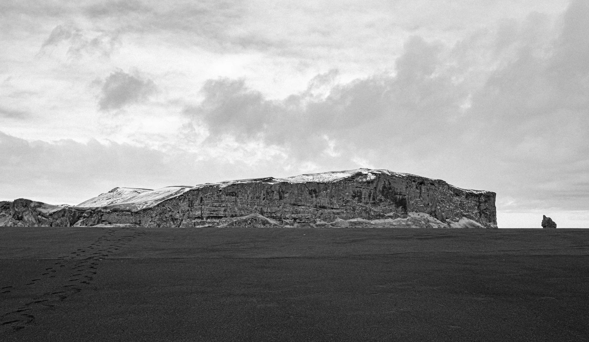 Black and white: a flat-topped headland above black sand, footprints trailing off to the left
