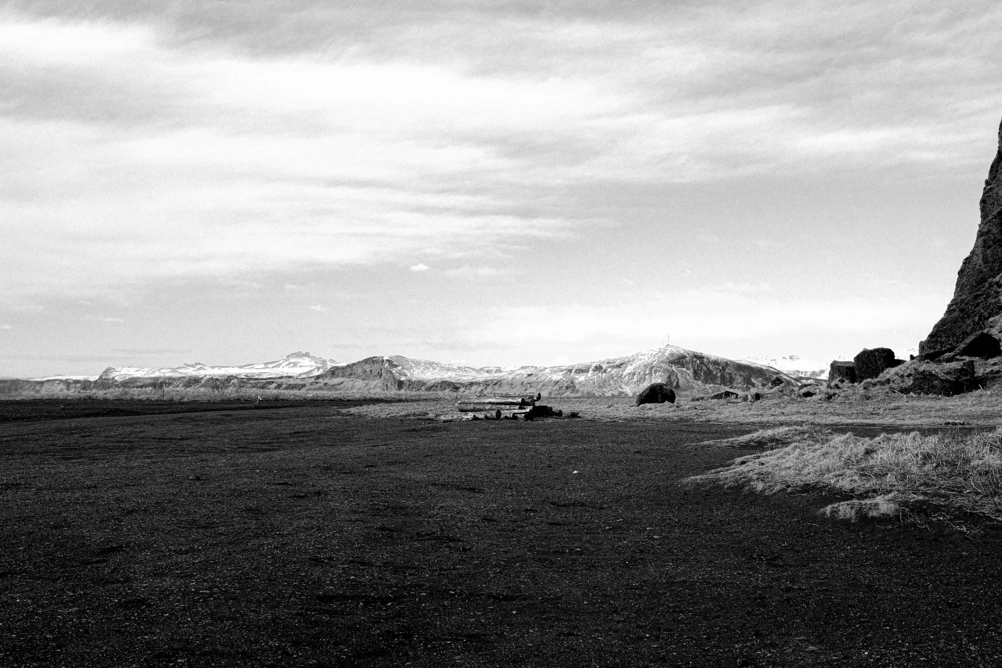 Black and white: a wide plain of dark gravel and dry grass, snowy mountains beyond and a cliff edge at right