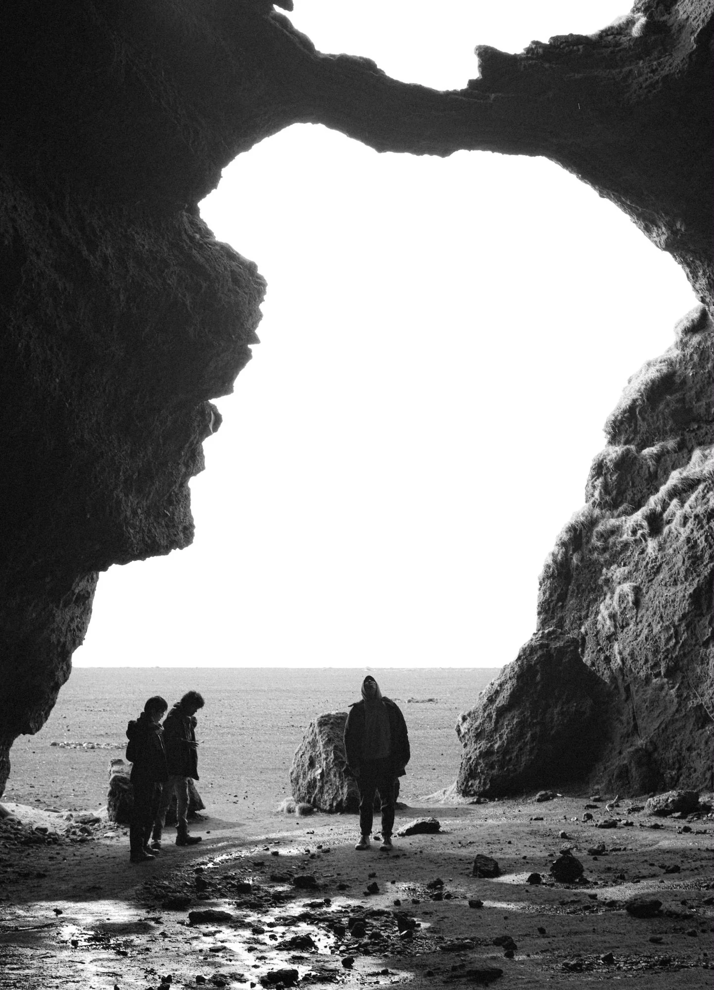 Black and white: three people standing at the mouth of a sea cave, silhouetted against a white sky